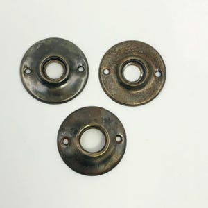 May include: Three antique, circular metal door hardware pieces. Each piece has a central hole and two smaller holes for screws. The metal has a tarnished, aged appearance with variations in color from dark gray to bronze.