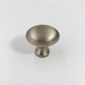 Op de afbeelding: Een kastknop van geborsteld nikkel met een ovaalvormige kop, een glad, afgerond bovenoppervlak en een taps toelopende cilindrische voet. Het metalen beslag heeft een zachte, matte afwerking met een zichtbare horizontale korrelstructuur.