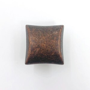 May include: A square cabinet knob with a dark, weathered copper finish. The surface features a mottled, textured appearance with varying shades of brown and bronze, and the edges of the knob curve slightly inward toward the center point.