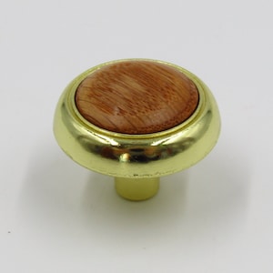 May include: A gold cabinet knob with a round wooden insert. The wood has a natural grain pattern.