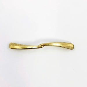 Könnte beinhalten: Eine goldfarbene Haarspange aus Metall mit geschwungener, länglicher Form. Die Oberfläche weist in der Mitte ein glattes, poliertes Finish auf, das an beiden abgerundeten Enden des Accessoires in ein körniges, texturiertes Muster übergeht.