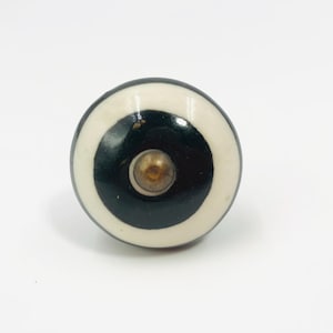 May include: A round cabinet knob featuring a concentric design with a cream-colored outer ring, a thick black middle band, and a small metallic center point. The surface is smooth and glossy, showcasing a clear contrast between the dark and light tones.