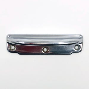 Puede incluir: Un tirador de armario de metal cromado con tres orificios de montaje circulares. El tirador rectangular tiene una parte superior curvada y una superficie reflectante. El herraje está diseñado para cajones o armarios.