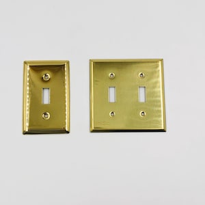 May include: Two polished brass wall plates with a reflective finish. One plate is rectangular with a single vertical toggle switch opening and two screw holes. The other is a square plate with two vertical toggle switch openings and four screw holes.