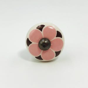 May include: A round cabinet knob with a cream-colored base featuring a pink six-petal flower design. The center of the flower has a dark metallic circular accent, and dark brown triangular shapes are positioned between each of the pink petals.