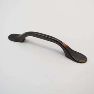 Puede incluir: Un tirador de armario negro con un diseño curvo. El tirador tiene un acabado liso y ligeramente redondeado.