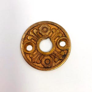 May include: A circular, antique brass-colored door plate with an ornate floral design. The plate has a central hole and two smaller holes, with a detailed pattern of flowers and curved lines radiating outwards.