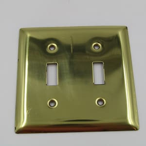 Könnte beinhalten: Eine goldfarbene Metall-Lichtschalterplatte mit zwei Kippschaltern und zwei Schraubenlöchern.