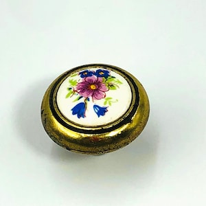 May include: A round cabinet knob featuring a metallic brass-toned finish with a weathered texture. The center is an inset white ceramic disc decorated with a floral pattern consisting of a central purple flower and smaller blue bell-shaped flowers.