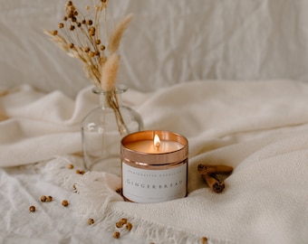 Pode incluir: Um pote de vela de cobre com um rótulo branco que diz "Handcrafted Candles Co. Gingerbread" e uma vela acesa dentro. A vela está sentada em uma manta branca com paus de canela e flores secas em um vaso de vidro.