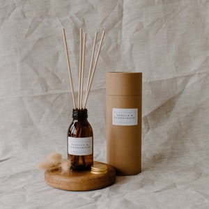 May include: A brown glass bottle with a white label that reads "Vanilla & Sandalwood" diffuser reeds. The bottle is sitting on a wooden round base with a gold metal lid. The bottle is next to a brown cardboard tube with a white label that reads "Vanilla & Sandalwood".