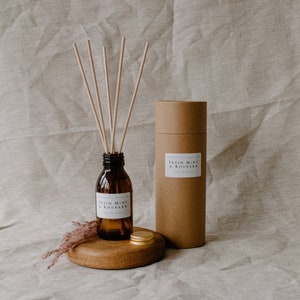 May include: A brown glass bottle with a white label that reads "Fresh Mint & Rhubarb" and a gold lid. The bottle is filled with a clear liquid and has wooden sticks sticking out of the top. The bottle is sitting on a wooden round base with a gold lid. The bottle is next to a brown cardboard tube with a white label that reads "Fresh Mint & Rhubarb".