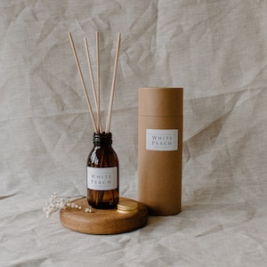 May include: A brown glass bottle with a white label that reads "White Peach" diffuser oil. The bottle is on a wooden round stand with a gold lid. There are wooden sticks in the bottle and a brown cardboard tube with a white label that reads "White Peach" diffuser oil.