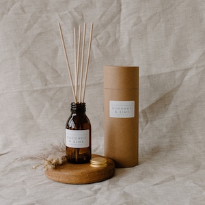 May include: A brown glass bottle with a diffuser stick set. The bottle has a white label with the text "Coconut & Lime" on it. The diffuser sticks are in a brown cardboard tube with the same label. The set is on a wooden surface with a gold lid.