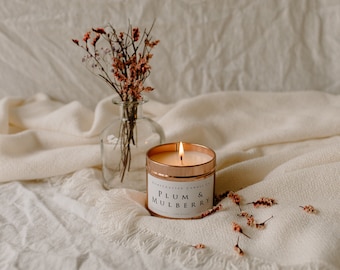May include: A lit candle in a copper tin with the label "Plum & Mulberry" on a white fabric background. Dried flowers in a glass vase are to the left of the candle.