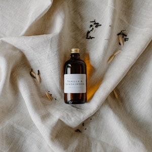May include: A brown glass bottle with a gold lid and a white label that reads "Vanilla & Sandalwood". The bottle is sitting on a light beige fabric surface.