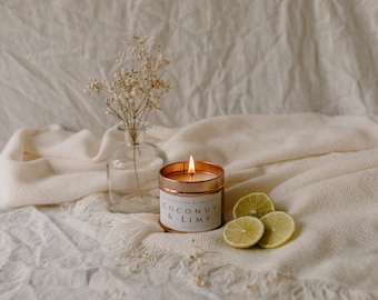 May include: A lit, copper-colored candle with the text "Coconut & Lime" sits next to lime slices and a small vase of dried flowers. The candle is in a round tin, and the flame is visible.