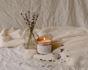 May include: A copper candle tin with a white label that reads "French Lavender" on a white fabric background with dried lavender sprigs in a glass vase.