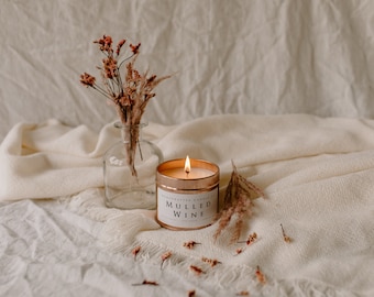 Pode incluir: Uma vela acesa, de cor dourada, com a etiqueta "Mulled Wine", está colocada ao lado de um pequeno vaso de vidro com flores secas. A vela está numa lata redonda com uma etiqueta branca. O fundo é um tecido creme.