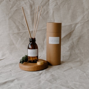 May include: A brown glass bottle with a gold lid and a white label that reads "Lime Basil & Mandarin" is on a wooden round base with a few sticks sticking out of the bottle. A brown cardboard tube with a white label that reads "Lime Basil & Mandarin" is next to the bottle.