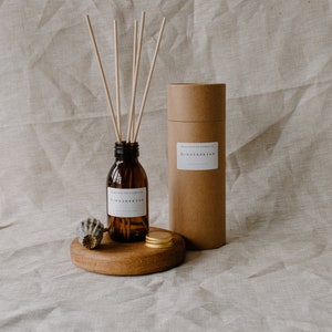 May include: A brown glass bottle with a white label that reads "Gingerbread" and a brown cardboard tube with a white label that reads "Gingerbread". The bottle is filled with a clear liquid and has a set of wooden sticks in it. The bottle and tube are sitting on a wooden surface.