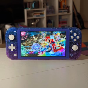 Può includere: Una console di gioco portatile con una scocca sfumata blu e viola glitterata. Lo schermo mostra la schermata del titolo di Mario Kart 8 Deluxe, con Mario su un kart rosso, la Principessa Peach e una pista da corsa colorata con fuochi d'artificio.