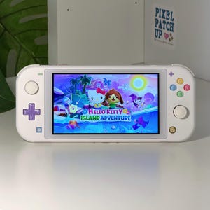 Puede incluir: Una consola de juegos portátil blanca con un mando de dirección morado y botones de acción coloridos. La pantalla muestra el título del juego Hello Kitty Island Adventure con personajes animados en un entorno tropical.