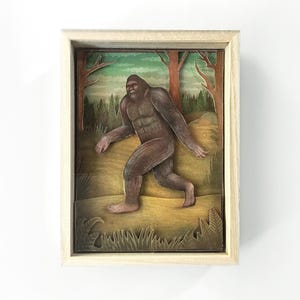 Peut inclure: Une œuvre d'art encadrée en papier découpé représentant un Bigfoot brun marchant dans une forêt. L'œuvre est encadrée dans un cadre en bois brun clair, avec un arrière-plan d'arbres et un paysage vert et jaune.