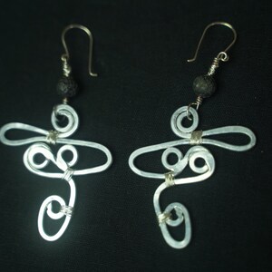 May include: A pair of silver wire earrings with a black lava rock bead at the top of each earring. The earrings are shaped like a stylized cross with a swirl design.