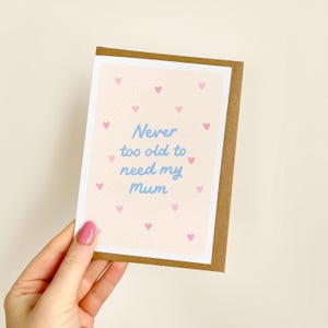 May include: A greeting card with a light brown border and a light pink background scattered with small pink hearts. The card features the blue text "Never too old to need my mum."