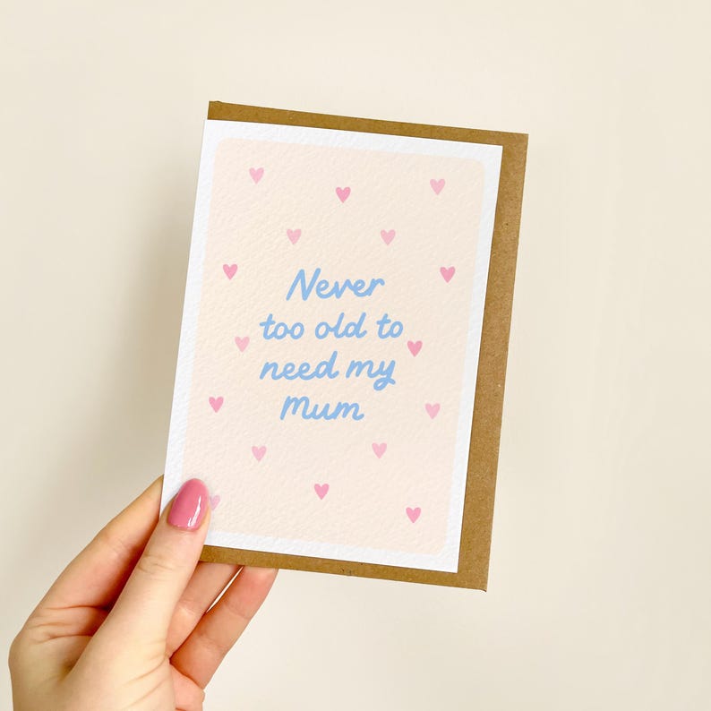 May include: A greeting card with a light brown border and a light pink background scattered with small pink hearts. The card features the blue text "Never too old to need my mum."