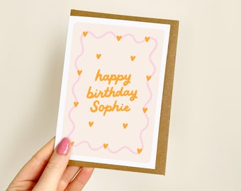 May include: A birthday card with a brown border and a light peach background. The card features a pink wavy border with orange hearts and the words "happy birthday Sophie" in orange script.