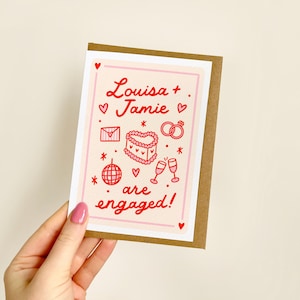 May include: An engagement card with a pink border and a brown backing. The card features red text that reads "Louisa + Jamie" and "are engaged!" with illustrations of hearts, rings, a cake, and champagne glasses.