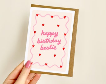 May include: A greeting card with a brown border and a light pink background. The card features the text "happy birthday bestie" in pink, surrounded by a wavy pink line and small red hearts. The card is held by a hand.