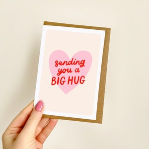 May include: A greeting card with a pink heart and the words "sending you a BIG HUG" in red. The card has a white border and a brown paper backing. The background is a light beige colour.