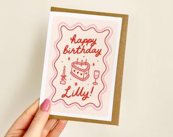 May include: A birthday card with a brown border and a light pink background. The card features the words "happy birthday Lilly!" in red script, along with illustrations of a cake, candle, and wine glass.