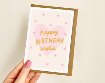 May include: A greeting card with a light pink background and a large pink heart in the center. The words "happy BIRTHDAY bestie" are written in yellow. Small pink hearts are scattered around the card. The card has a brown border.