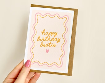May include: A greeting card with a brown border and a light pink background. The card features the text "happy birthday bestie" in gold script, framed by wavy pink and gold lines, and a small pink heart.