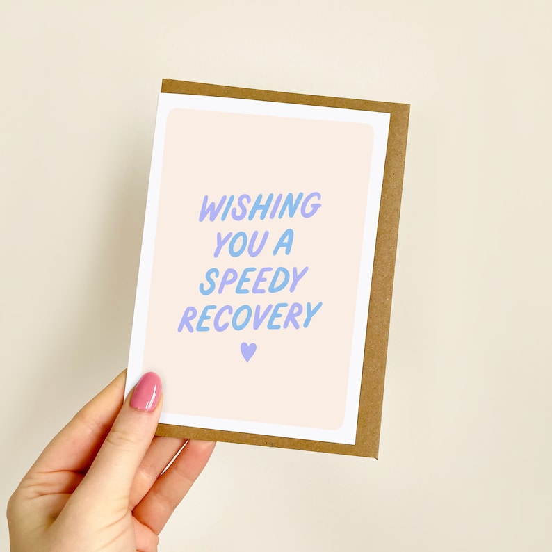 May include: A greeting card with the text "WISHING YOU A SPEEDY RECOVERY" in blue and purple gradient lettering. The card has a light peach background with a small purple heart. The card is in a brown paper frame.