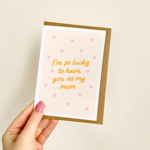 May include: A greeting card with a light pink background and small pink hearts. The card has the text "I'm so lucky to have you as my mum" in orange script. The card is held by a hand with pink nail polish.