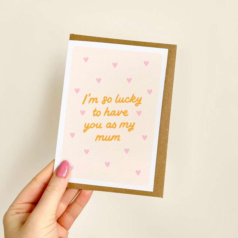 May include: A greeting card with a light pink background and small pink hearts. The card has the text "I'm so lucky to have you as my mum" in orange script. The card is held by a hand with pink nail polish.