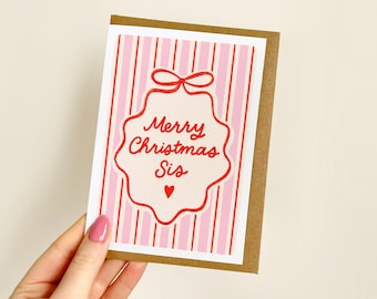 May include: A rectangular greeting card with a brown kraft paper border. The front features a pink and white vertical striped background with a central white scalloped ornament shape outlined in red, containing the red text Merry Christmas Sis.