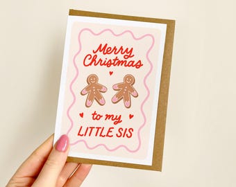 May include: A rectangular greeting card with a light pink background and a wavy pink border. The text reads Merry Christmas to my LITTLE SIS in red. Two brown gingerbread figures with pink icing details are centred with small red hearts.