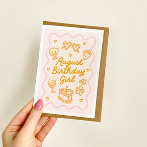 May include: A greeting card with a brown border and a pink and white interior. The card features the text "August Birthday Girl" in gold, along with illustrations of hearts, sunglasses, a seashell, a strawberry, a flower, a cake, and a cocktail glass.