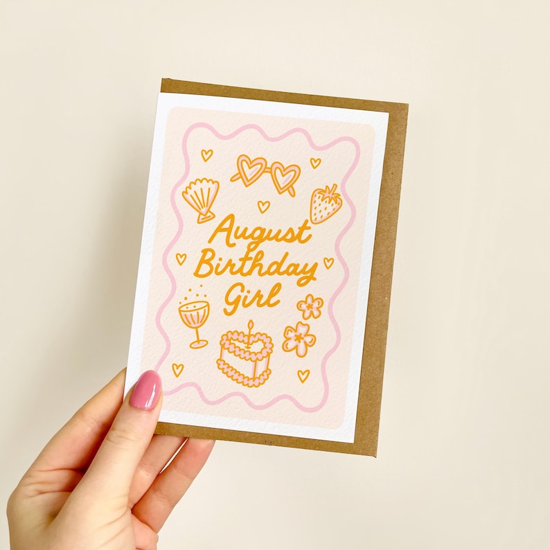 May include: A greeting card with a brown border and a pink and white interior. The card features the text "August Birthday Girl" in gold, along with illustrations of hearts, sunglasses, a seashell, a strawberry, a flower, a cake, and a cocktail glass.