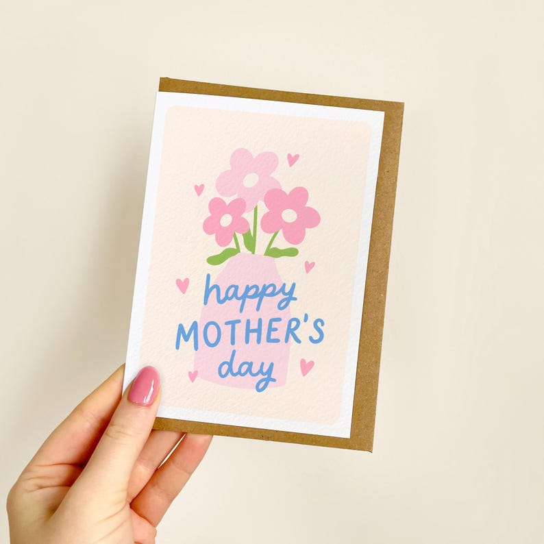 May include: A Mother's Day greeting card featuring a pink vase with pink flowers and green leaves. The card has the text "happy MOTHER'S day" in blue. Small pink hearts are scattered around the design. The card is held by a person.