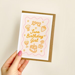 K&ouml;nnte beinhalten: Eine Geburtstagskarte mit braunem Rand wird von einer Hand gehalten. Die Karte hat einen rosa Hintergrund mit orangefarbenen Illustrationen von Sonnenbrillen, einer Muschel, einer Erdbeere, einem Kuchen, Blumen und einem Cocktailglas. Der Text lautet "June Birthday Girl."