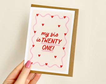 May include: A birthday card with a brown border and a light beige background. The card features a pink wavy border with red hearts and the text "my sis is TWENTY ONE!" in red.