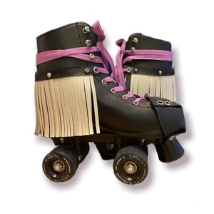 May include: Black roller skates with lilac laces and fringe detailing. The skates have black wheels and a black toe stop. The skates have silver rivets and a lilac strap around the ankle.