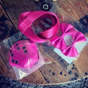 Peut inclure: Ensemble d'accessoires roses, comprenant une sangle enroulée, une paire d'ornements en forme de nœud papillon et un article carré avec des œillets métalliques. Les articles sont emballés dans du plastique transparent et disposés sur une surface en bois.